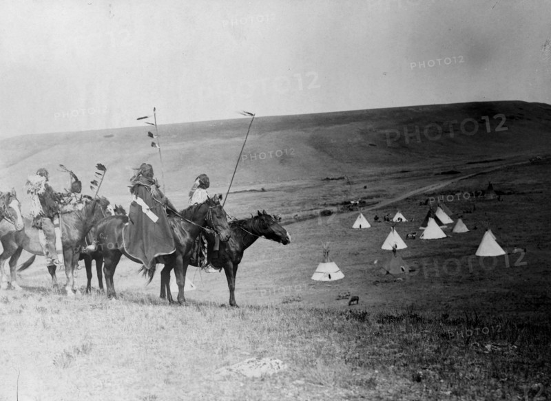 Four Atsina Indians on horseback overlooking tepees in valley beyond