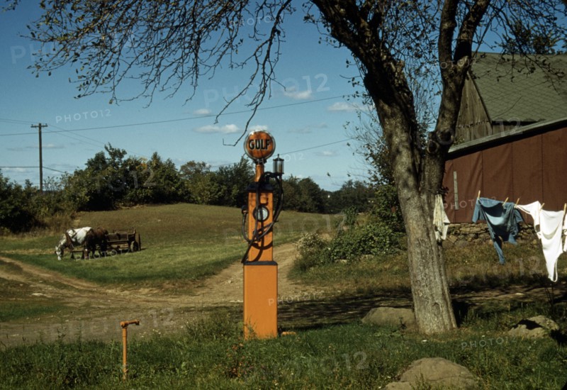 Gas pump with clothesline, barn and horse-drawn wagon in background.