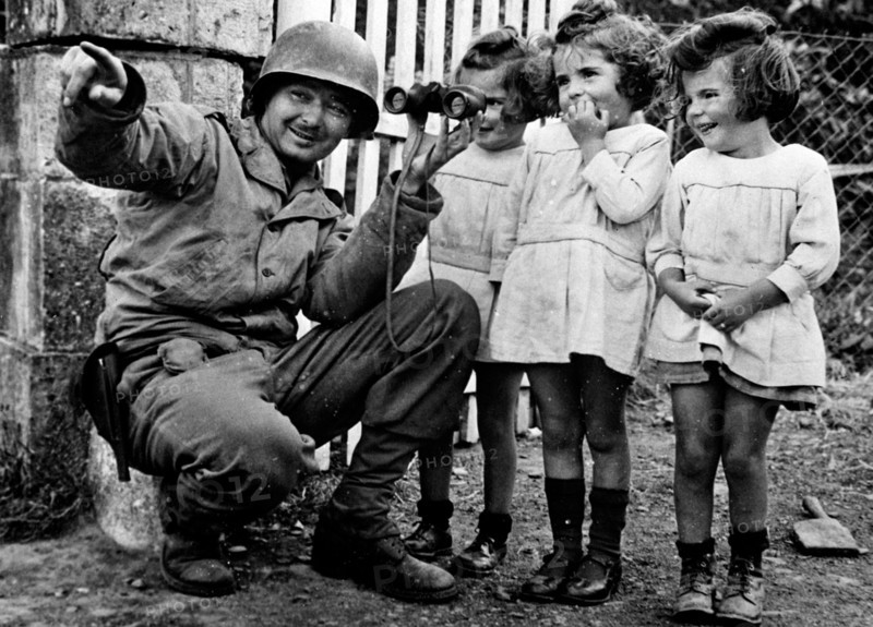 Soldier holding binoculars up to three little girls.