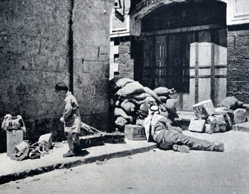 Photograph of an Arab soldier guarding a road block in the Old City in Jerusalem, whilst a young boy searches through the rubble