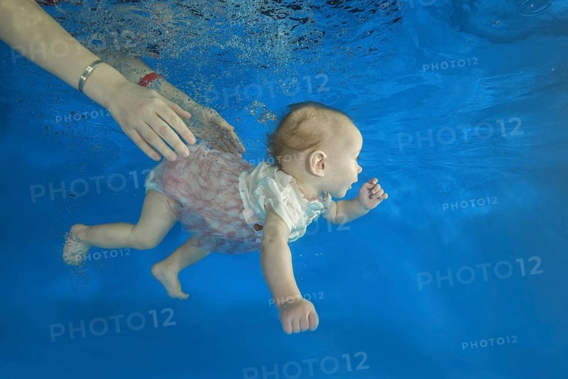 Infant girl dives in a pool with hands of her mother holding it