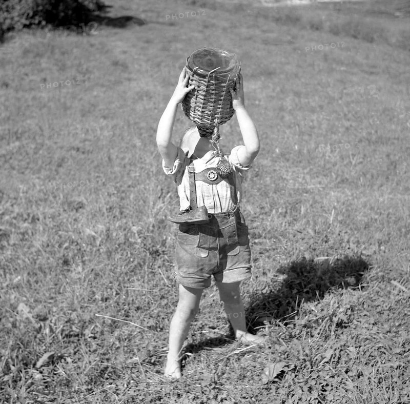 Child stands barefoot in a meadow and drinks water from a wooden-coated vessel
