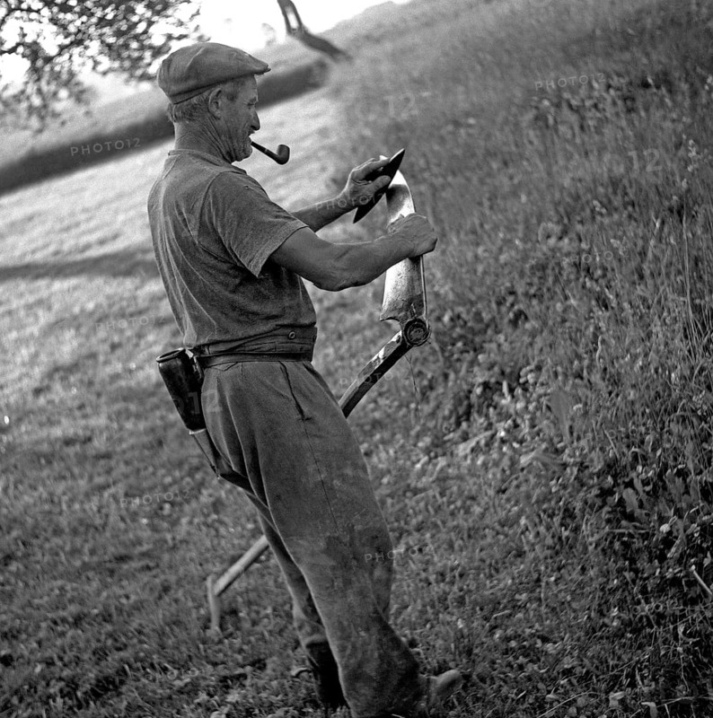 Man with a pipe sharpened in his mouth sharpens a scythe with the help of a grindstone
