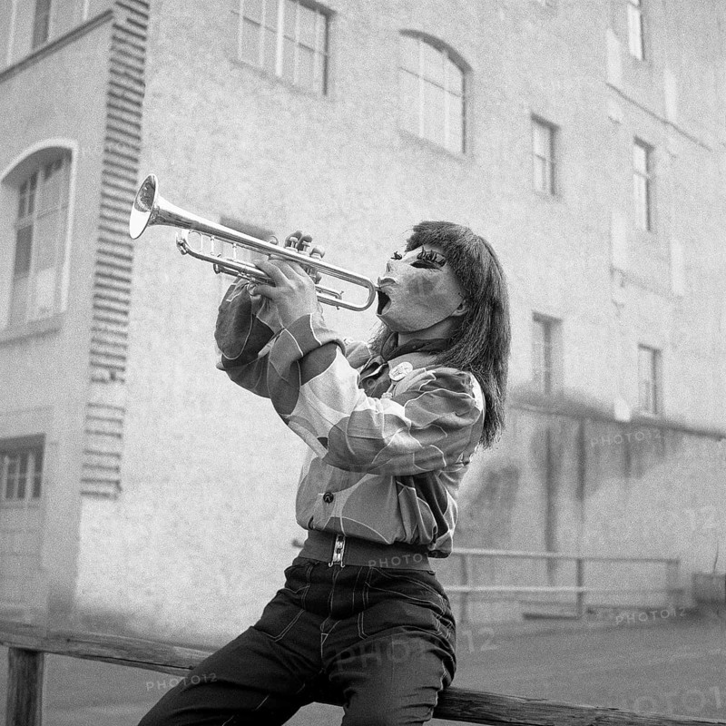 Man with shirt and mask sits on a wooden railing and plays trumpet