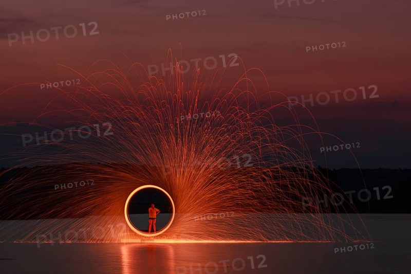 A man stands on a platform in a lake and creates a hoop of fire with glowing steel wool around him