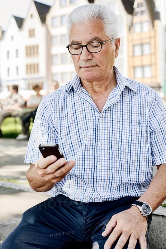 Grey-haired senior sits on a wall with his smartphone in his hand