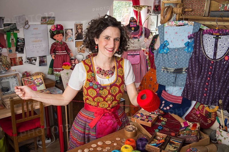 Button maker in colorful dirndl with Collier button trimmings in handicraft studio working with red yarn and utensils for Posamentenknopfe