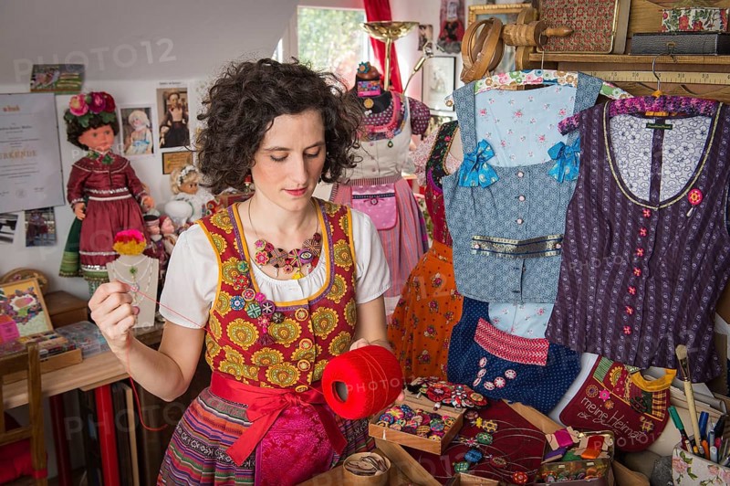 Button maker in colorful dirndl with Collier button trimmings in handicraft studio working with red yarn and utensils for Posamentenknopfe