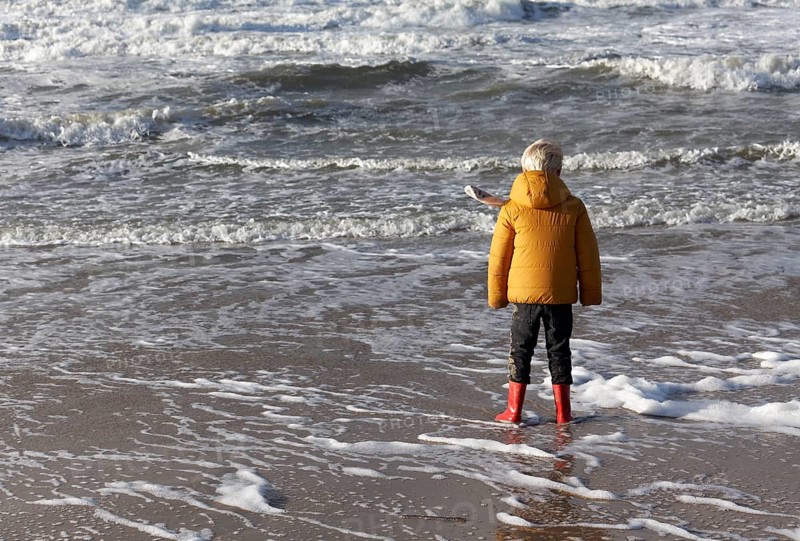 Little boy in winter clothes standing in the surf on the beach