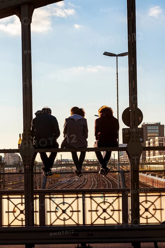 Three youths sitting on the balustrade of the Hackerbrucke bridge over the railway tracks looking into the distance