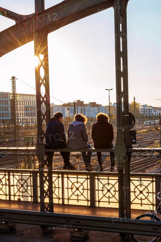 Three youths sitting on the balustrade of the Hackerbrucke bridge over the railway tracks looking into the distance