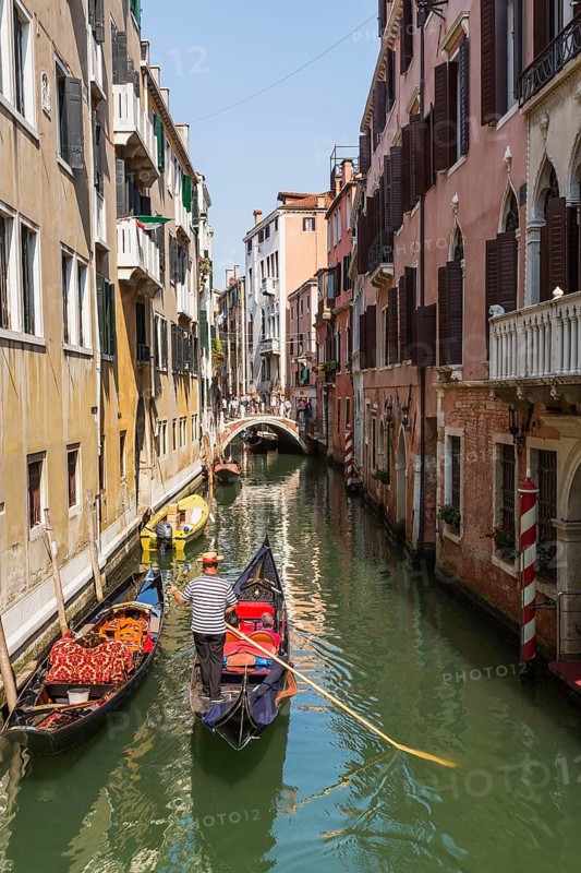Gondolier steering gondola with tourists on narrow canal with historical buildings