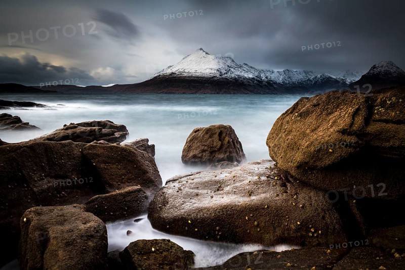Big rocks in the water of the North Sea with snow-covered Cullin mountains in the background