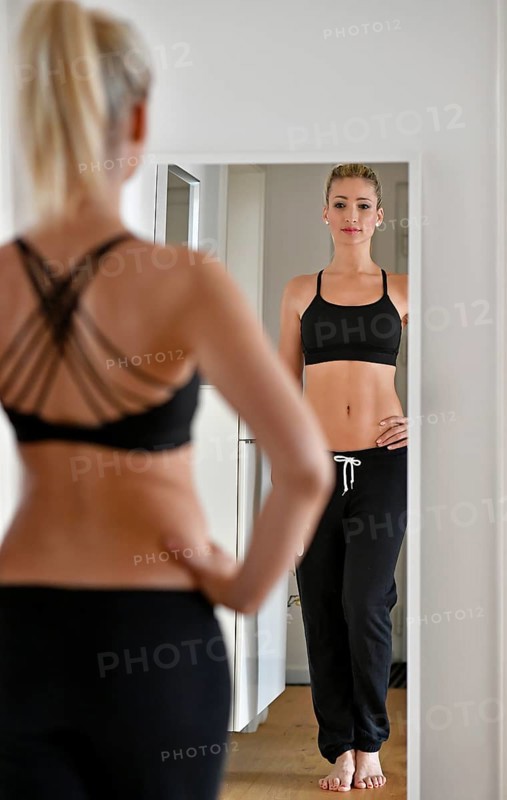Young woman with sports bra and sweatpants looks at herself in the mirror