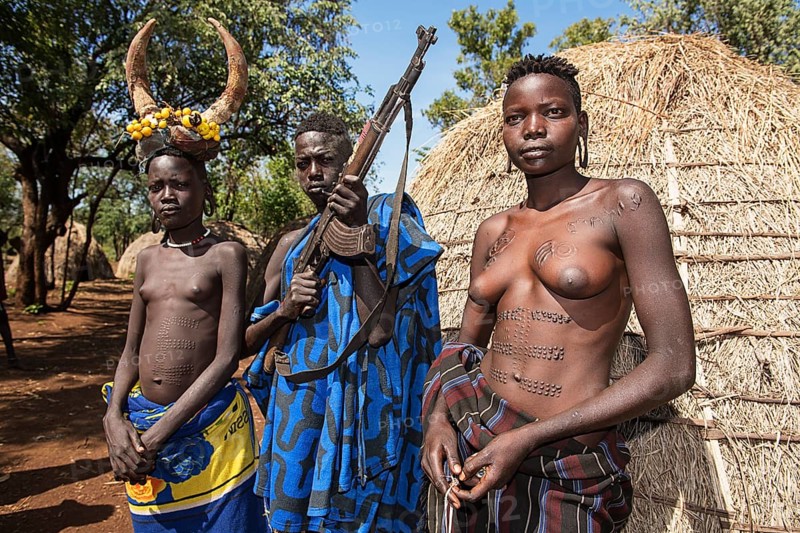 Young girls with decorative scars and man with Kalashnikov in front of hut