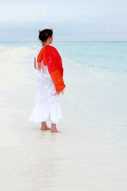 Woman with a red scarf standing on a white beach looking at the turquoise blue sea