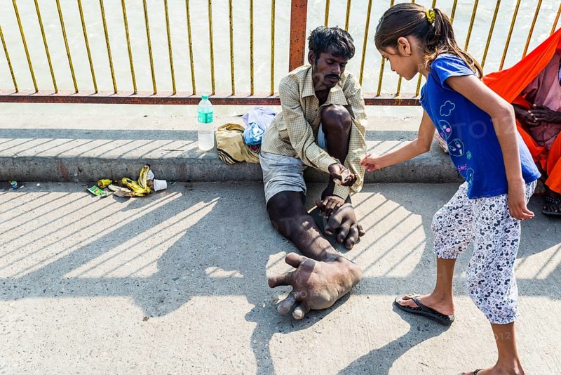 A beggar with Elephantiasis at his feet is receiving some money from a girl at Harki Pauri Ghat at the holy river Ganges