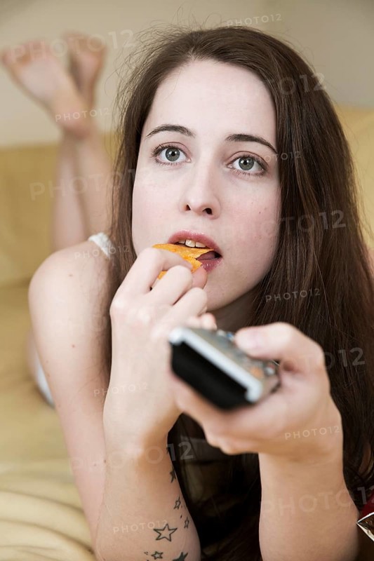 Young woman watching TV on the couch with chips and the remote control in hand