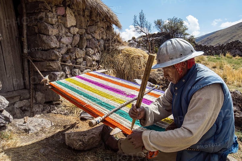Old Indio man with helmet weaving a colorful rug with a simple loom