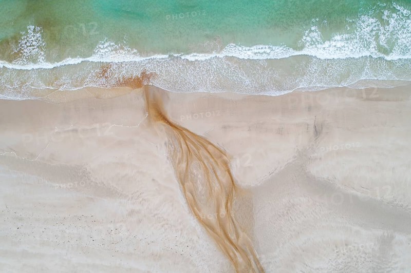 Creek with reddish ferruginous water flows over a sandy beach into the sea