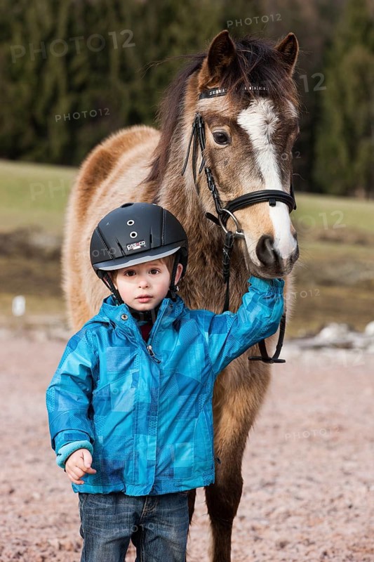 Young child wearing a riding helmet standing beside a pony