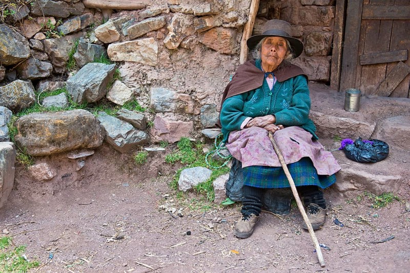 Mature Quechua Indian woman wearing a hat sitting on a stone in front of a doorway