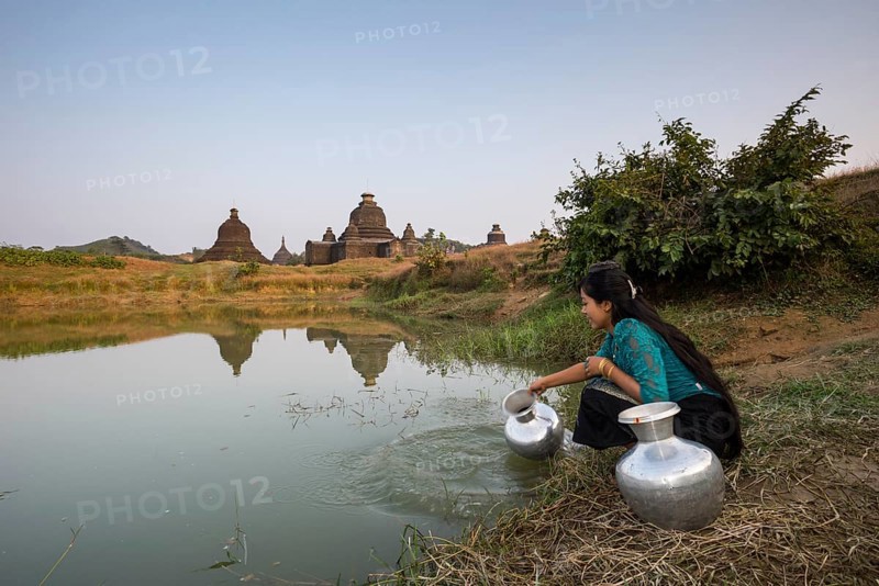Smiling girl drawing water from a water hole with a vessel made of aluminium