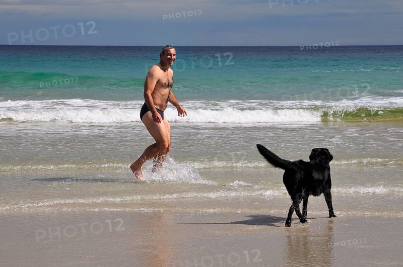 Man in swimming trunks playing with a dog on the beach of Rispond Bay