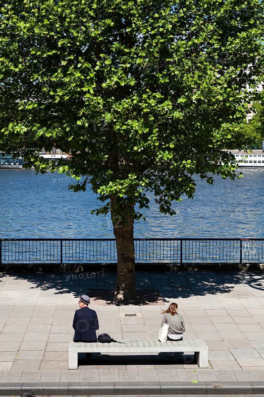 Two people sitting on concrete bench by river with tree