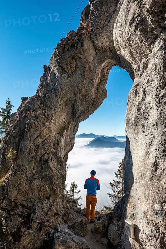 Young man stands in the rock gate Breitensteinfensterl with a view of the high fog above the valley