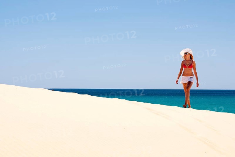 Woman wearing a hat and a bikini walking along a sandy beach