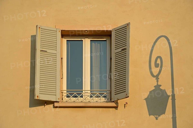 A facade in Ajaccio with a lone window and the shadow of a street lamp illuminated by warm morning light. Ajaccio is the capital of the mediterranean island of Corsica