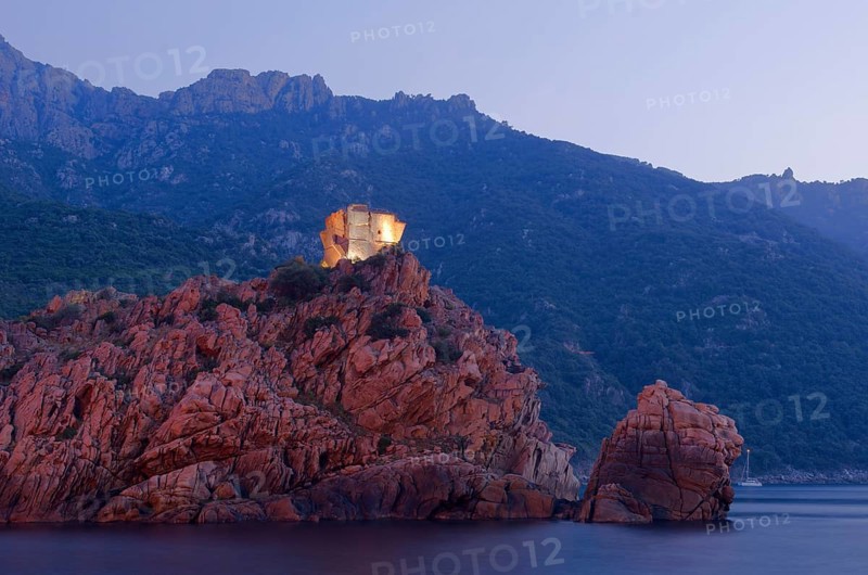 The flood lit Genoese Tower of Porto on top of a rocky hill and the Gulf of Porto at the blue hour