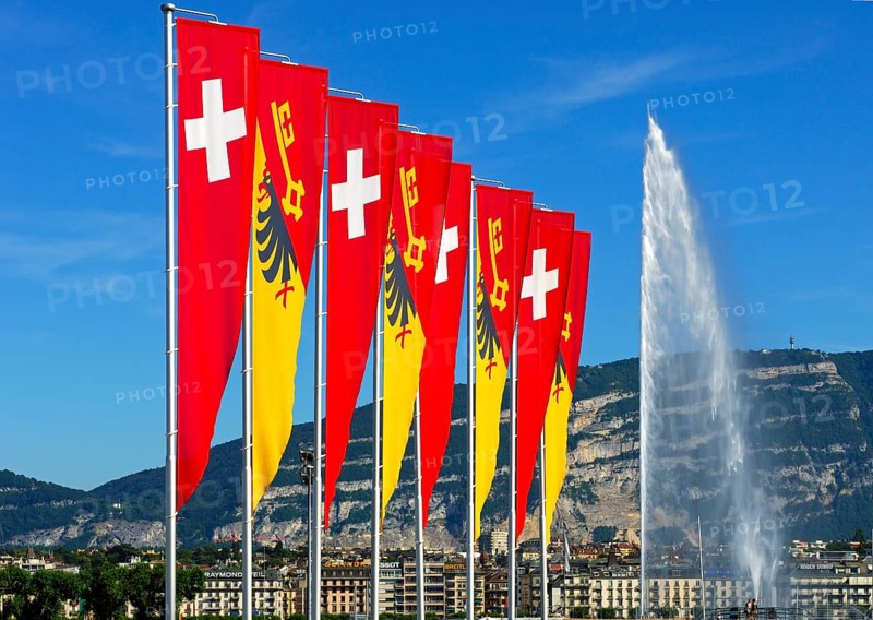Flags of the Swiss Confederation and the Canton of Geneva on the promenade of Lake Geneva with the Jet d'Eau fountain and the summit of Saleve Mountain at the rear