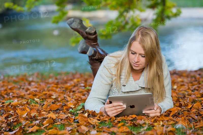 Young woman lying on her belly on the ground in a park and looking at a tablet PC