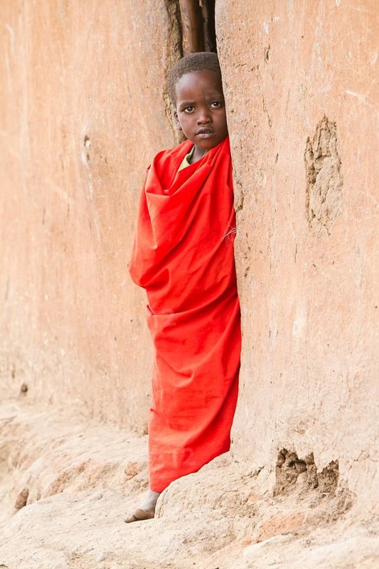 Maasai child wearing traditional dress looking out of a mud hut