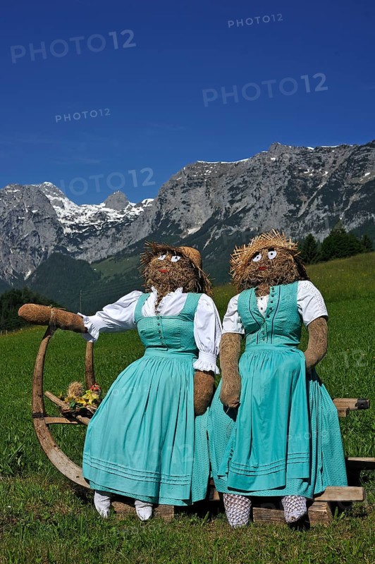 Two straw dolls or figures in dirndl dresses sitting on a sledge with long horn-shaped runners on a meadow
