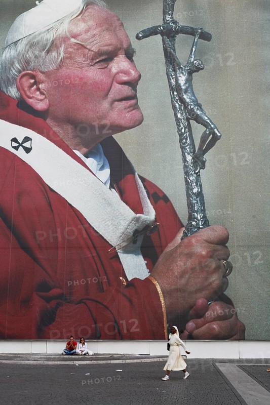 Two tourists sitting in front of a huge poster of Pope John Paul II