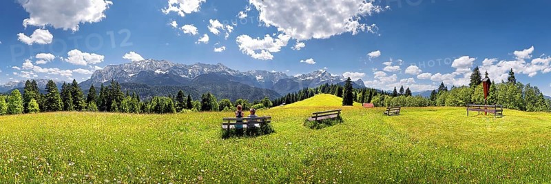 Hikers relaxing on a bench in the middle of a flowery meadow with panoramic views towards the Wetterstein Massif with the Alps and Zugspitze Mountain