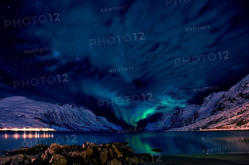 Aurora over a fjord with snow-covered mountains and an illuminated village