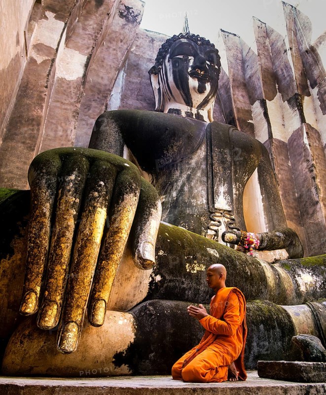 Praying monk neeling in front of the hand of the Buddha statue Phra Achana which is decorated with gold leaf
