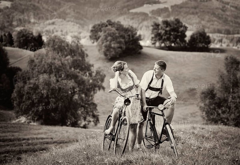 Man wearing leather pants and a woman wearing a dirndl on old bicycles within a natural landscape