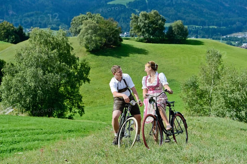 Man wearing leather pants and a woman wearing a dirndl on old bicycles within a natural landscape