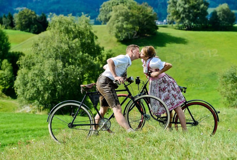 Man wearing leather pants and a woman wearing a dirndl kissing on old bicycles within a natural landscape