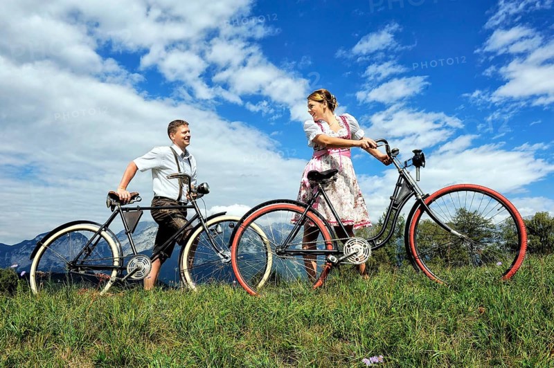 Man wearing leather pants and a woman wearing a dirndl with old bicycles within a natural landscape