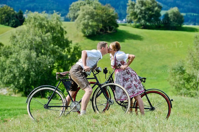 Man wearing leather pants and a woman wearing a dirndl kissing on old bicycles within a natural landscape