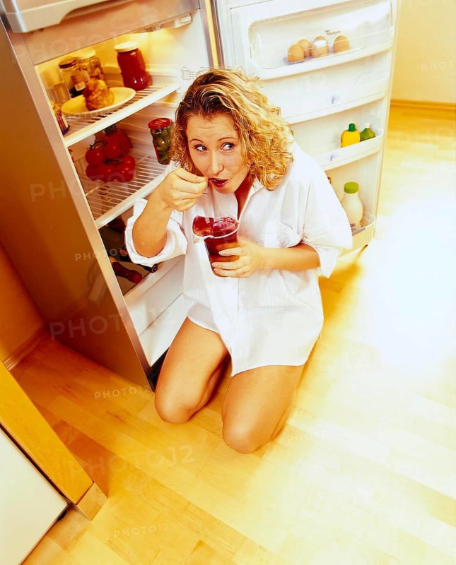 Woman kneeling in front of an open refrigerator in a nightshirt and eating red jelly