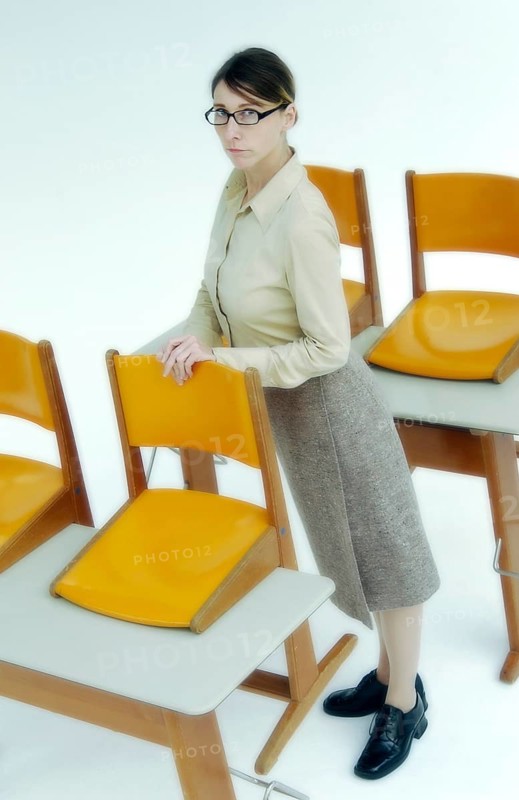 Strict female teacher standing at old school desks with orange chairs