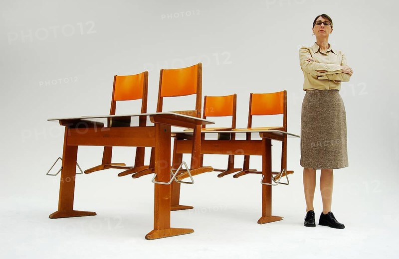 Strict female teacher standing next to old school desks with orange chairs