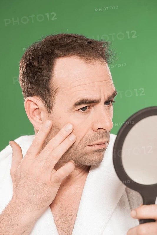 Man wearing a bathrobe observing himself critically in a hand mirror